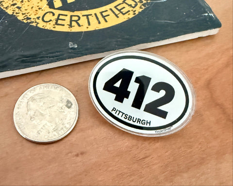 A circular white and black '412 Pittsburgh' sticker shown next to a US quarter for scale on a wooden tabletop, with a 'Yinzer Certified' book in the background.