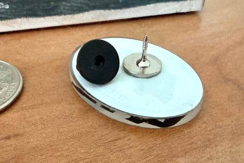 A close-up of a small oval metal earring or accessory with a black circular element and a silver post, placed on a wooden surface next to a coin for scale. A branded card reading Vinzer is partially visible in the background.