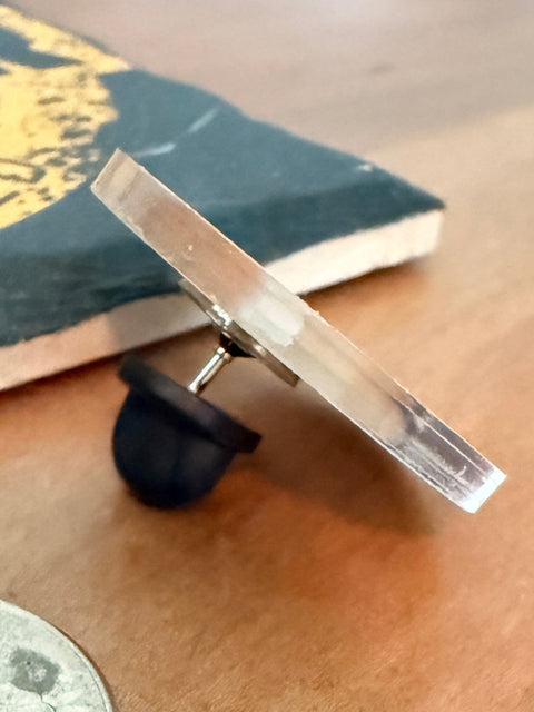 A small clear rectangular pin with a black rubber base is shown on a wooden tabletop next to a silver coin for scale. A partially visible book or notebook appears in the background.